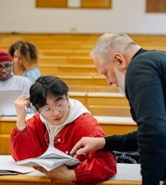 A teacher helps a student with coursework in a university lecture hall setting.