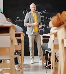 Bald teacher explaining geometry on blackboard to attentive students in classroom.