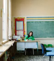 Female teacher writes notes at desk in a bright, spacious classroom with chalkboard.